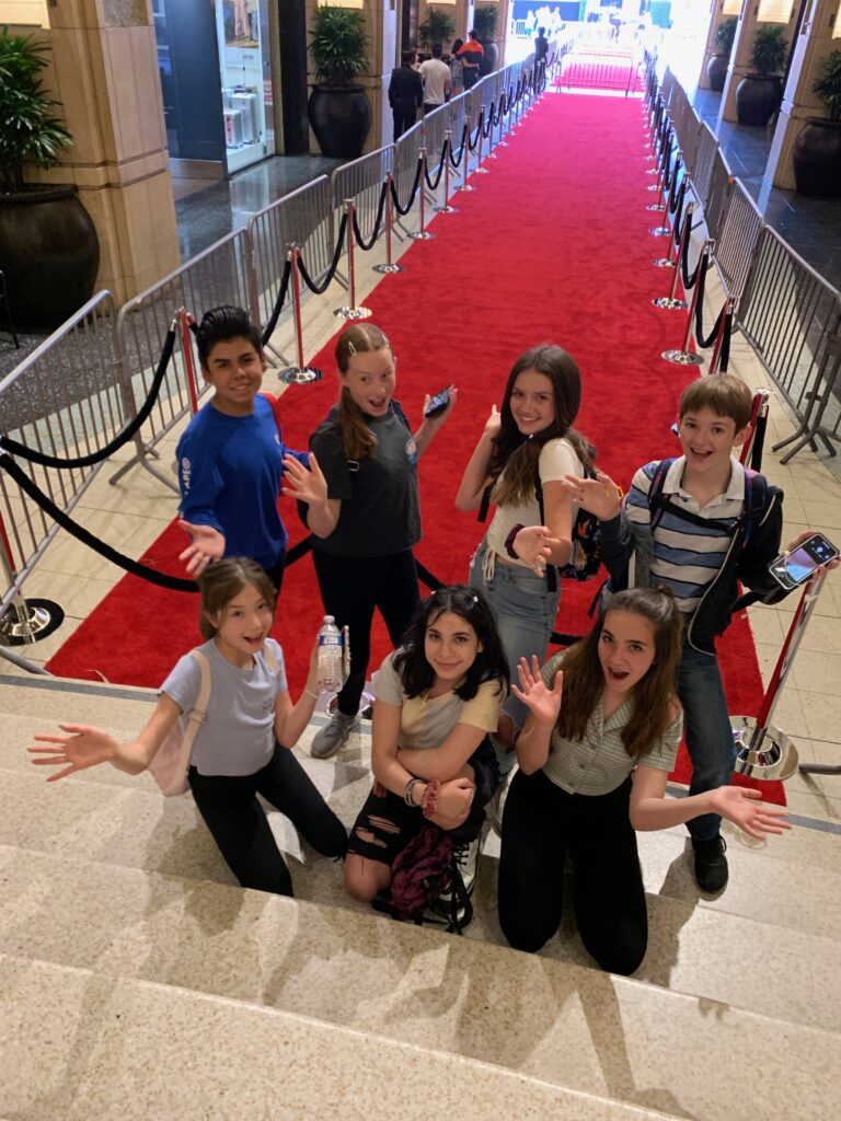 A group of children and teens smiling at the camera with a red carpet behind them